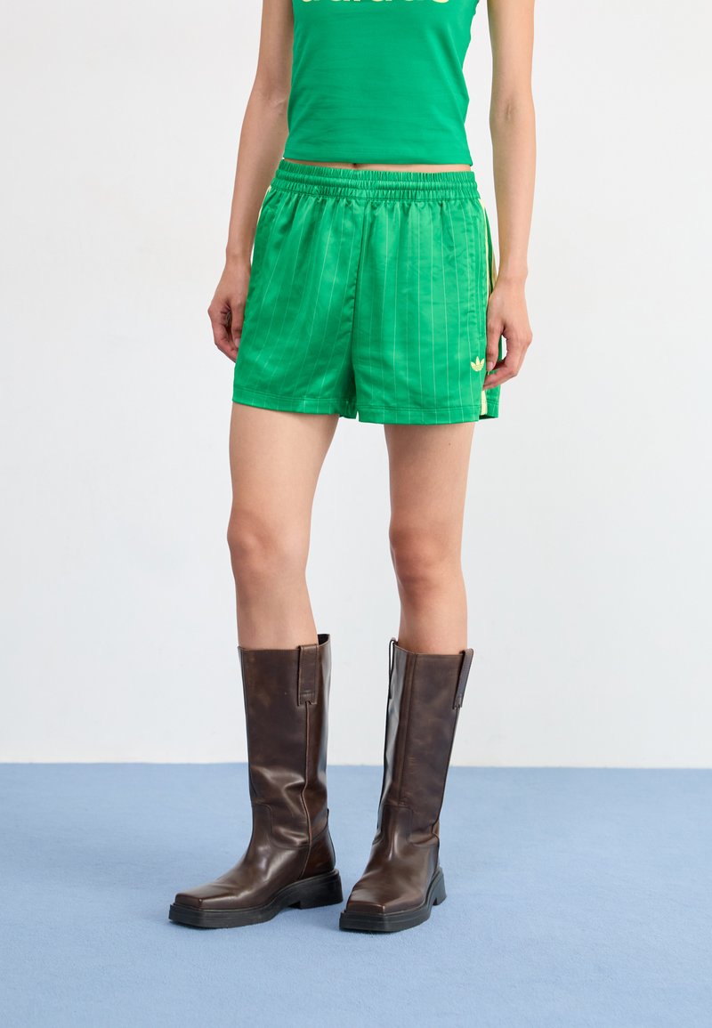 Green, striped shorts with an elastic waistband and a small logo on the hem, paired with tall, brown leather boots on a blue surface.