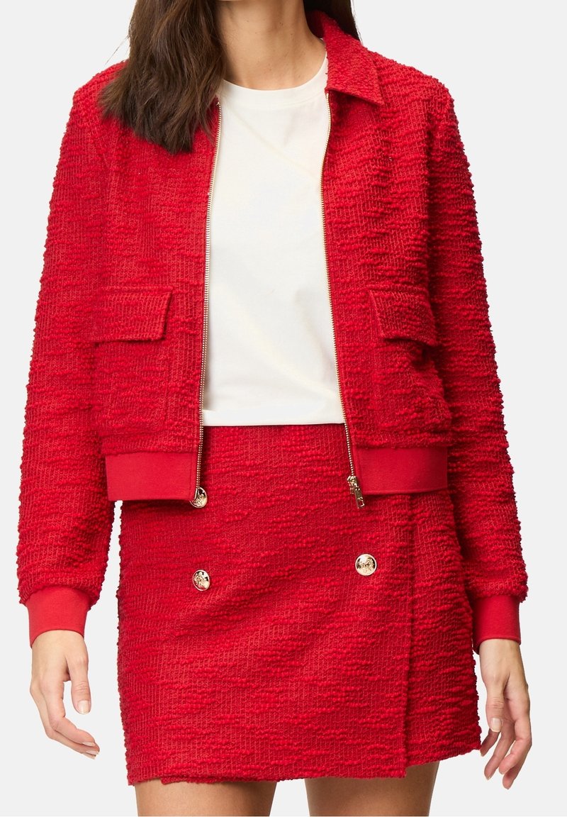 Veste zippée rouge texturée avec deux poches avant, associée à une jupe assortie ornée de boutons dorés. Les deux ont une coupe courte.