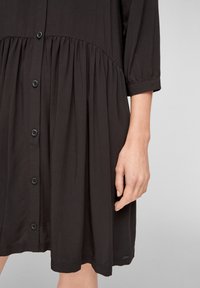 Robe noire boutonnée en tissu doux. Présente un corsage ajusté, une jupe plissée, des manches trois-quarts et des boutons sur le devant.