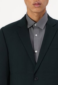 Dunkelgrüner Blazer mit Reverskragen, einreihiger Knopfverschluss und einer Brusttasche. Kombiniert mit einem hellgrauen Hemd mit Knöpfen.