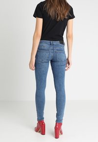 Úzké modré džíny z denimu s pasem ve střední výšce, dvěma zadními kapsami a hladkou strukturou. Doplněné červenými botami na podpatku.