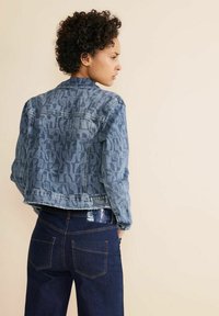 Chaqueta de denim con un tono azul desgastado, que presenta patrones oscuros abstractos, un corte corto y una etiqueta de marca en el dobladillo. Combinada con jeans oscuros.