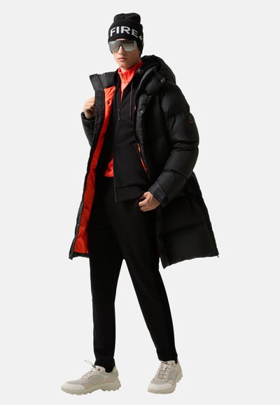 Mann trägt eine schwarze Steppjacke mit orangefarbener Fütterung, eine schwarze Mütze mit weißem Text, übergroße Sonnenbrille, schwarze Hosen und beige Sneakers.