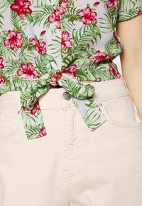 Camisa floral com hibiscos vermelhos num padrão de folhas de palmeira verdes, atada na cintura. Combinada com calças de ganga beige claro.