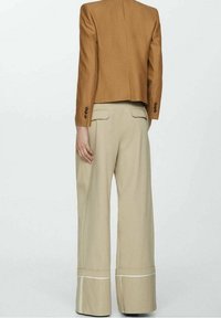 Hnědý zkrácený blejzr s dlouhými rukávy, spárovaný s béžovými kalhotami s širokými nohavicemi, které mají bílý lem na spodním okraji a vzadu kapsy.