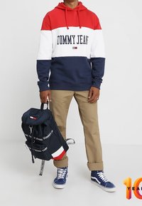 Felpa con cappuccio a blocchi di colore rosso, bianco e blu navy con logo "TOMMY JEANS"; pantaloni beige; zaino blu navy con dettaglio logo; scarpe da ginnastica blu.
