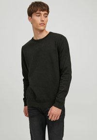 Donkergroene trui met een ronde hals, gestructureerde stof, ribgebreide manchetten en zoom. Draagt met donkere spijkerbroek, gedeeltelijk zichtbare handen.