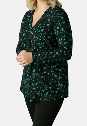 Vrouw draagt een zwarte blouse met lange mouwen en een blauw bloemenpatroon, staat met gevouwen handen voor een effen achtergrond.