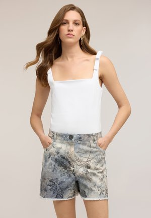 Femme aux longs cheveux ondulés portant un top blanc sans manches avec bretelles et un short en jean à fleurs taille haute, les mains dans les poches.