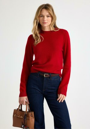 Kvinde med blond hår iført en rød sweater og mørkeblå jeans, der holder en brun læderhåndtaske mod en ensfarvet hvid baggrund.