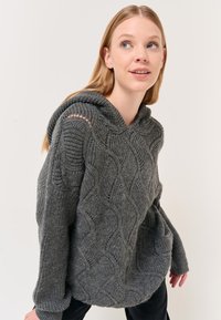 Felpa grigia a maglia con motivo a onda testurizzato, maniche ampie e vestibilità rilassata. Presenta un cappuccio con cordino e dettagli sulle spalle a giorno.