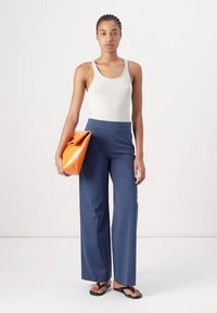 Pantalons larges navy associés à un débardeur blanc à côtes ; le mannequin tient une pochette structurée orange ; des tongs noires complètent la tenue.