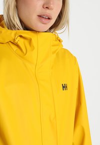 Gele waterdichte jas met een hoge kraag, verstelbare trekkoord en zwart logo op de borst. Gladde textuur en naadloos ontwerp.