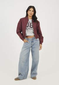 Veste bomber bordeaux, t-shirt gris court avec motif, jeans larges bleu clair, chaussures marron, design minimaliste.