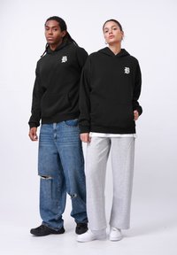 Două modele îmbrăcate în hanorace negre cu buzunar frontal și logo. Unul poartă blugi albaștri uzati, iar celălalt pantaloni de trening gri deschis și adidași albi.