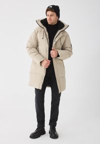 Vīrietis, tērpjoties bēšā pildītā garajā mētelī ar melnu rāvējslēdzēju, melnā turtleneck, melnās biksēs, melnās zābakos un melnā beanie uz baltā fona.