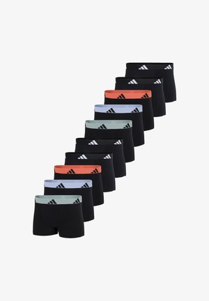 Junger Mann steht barfuß und trägt schwarze Boxershorts mit einem orangefarbenen Bund und einem schwarzen Logo vor einfachem hellem Hintergrund.