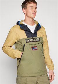 Gul, olivengrøn og sort letvægtsjakke med hætte, der har en frontlomme, lynlås og et norsk flag-patch.