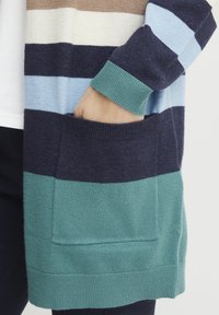 Krāsains kardigāns ar joslām, kas veidotas jūras laivā, tirkīza, gaiši zilā, krēmkrāsā un brūnā. Ietver priekšējo kabatu un ribotas piedurknes detaļām.