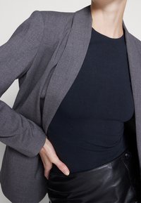 Blazer gris ajustado con un acabado texturizado, combinado con una blusa negra lisa. El conjunto incluye una falda de cuero negra con acentos de botones.