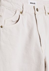 Jeans de denim bege com corte reto, bolsos frontais, ferragens em latão e um tecido texturizado. O rótulo diz "ROLLA'S."