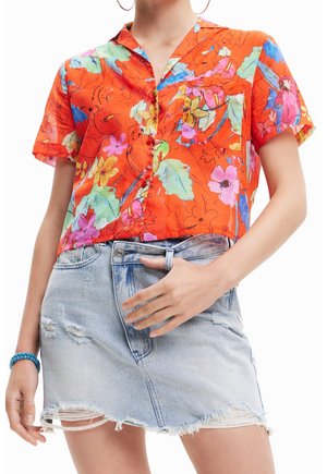 Femme portant un t-shirt orange vif à manches courtes avec des imprimés floraux et d'oiseaux et une jupe en jean bleu clair déchirée avec un ourlet effiloché.