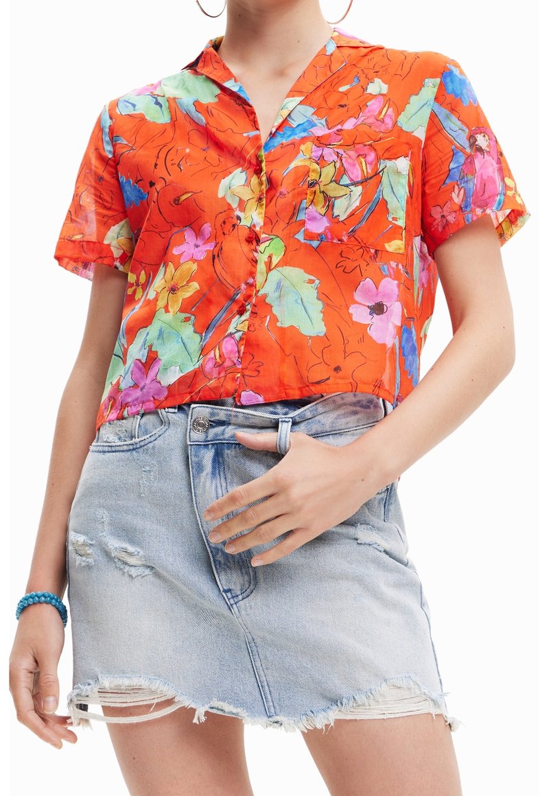 Femme portant un t-shirt orange vif à manches courtes avec des imprimés floraux et d'oiseaux et une jupe en jean bleu clair déchirée avec un ourlet effiloché.