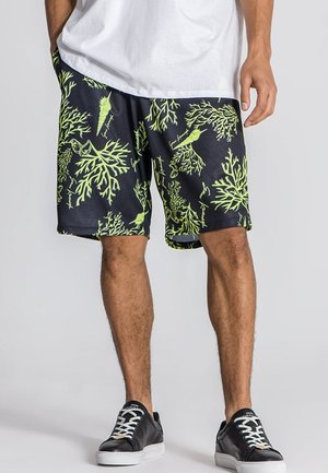 Mężczyzna ubrany w czarne spodenki z neonowozielonymi wzorami wodorostów i muszli, białą koszulkę oraz czarne buty sportowe z białymi podeszwami i sznurowadłami.