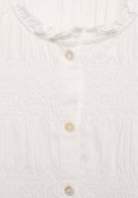 Biela blúzka s volánovým výstrihom, obsahujúca elastický smock, šesť gombíkov a ľahký materiál. Hladká textúra s jemným leskom.