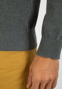 Grå strikket sweater med ribbede manchetter. Nærbillede af underarmen viser blød tekstur og tætsiddende design. Sammen med gule chinos.