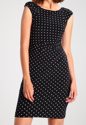 Frau trägt ein eng anliegendes schwarzes Kleid mit weißen Punkten, kurzen Ärmeln und einem gerafften Detail in der Taille.