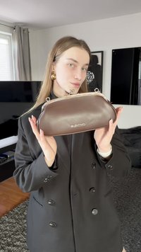 Brown leather clutch bag with silver hardware, rounded shape, and visible Valentino logo. Model holds bag and wears a black blazer.
