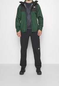 Veste imperméable verte et noire avec fermeture éclair, capuche ajustable et poignets élastiques, associée à un pantalon noir et des chaussures noires robustes.