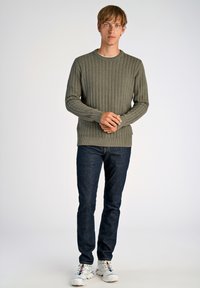 Pull en tricot vert avec un motif texturé, col rond et poignets côtelés, associé à un jean foncé et des baskets blanches.
