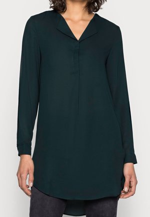 Vrouw draagt een donkergroen langärmelig tuniekblouse met een V-halskraag, gecombineerd met donkere broek, staand tegen een effen achtergrond.