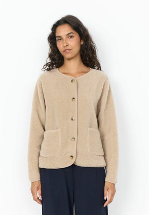Donna con capelli ricci che indossa una giacca in pile beige con bottoni e due tasche davanti, pantaloni scuri, in piedi su sfondo bianco.