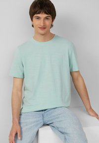 Licht muntgroen t-shirt met korte mouwen, gemaakt van zachte stof, met een ronde hals en een ontspannen pasvorm. G gedragen met lichtblauwe spijkerbroek.