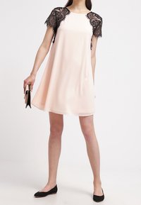 Pale pink sleeveless dress with black lace cap sleeves, flared shape, smooth fabric, and subtle sheen; paired with black flats and a clutch.
