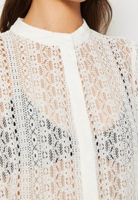 Vit blus med spetsknäppning, som har invecklade mönster, mandarin-krage och en genomskinlig textur.