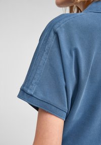 Krátký rukáv, modré polo tričko s texturovanou tkaninou. Obsahuje skládací límec a viditelné stehy na rukávech, což zdůrazňuje čistý design.