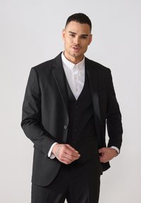 Schwarze Sakkoblazer mit Reverskragen, taillierter Passform und Ein-Knopf-Verschluss. Darunter ein weißes Hemd. Glattes Gewebe, keine sichtbaren Muster.