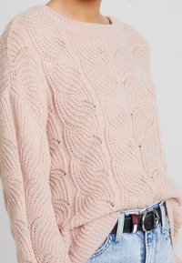 Ljust rosa kabelstickad tröja med rund halsringning och lös passform. Bärs över högmidjade jeans med ett svart bälte. Mönsterdetalj med textur.