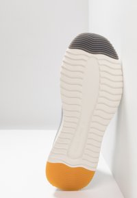 Sole di scarpa atletica grigia con accenti bianchi e arancioni. Presenta un motivo di battistrada ondulato e una texture liscia. Design arrotondato e a profilo basso.
