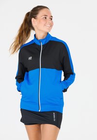 Kvinnors träningsjacka i blått och svart. Har dragkedja framtill, hög krage och sidofickor. Tillverkad av lätt, andningsbart material.