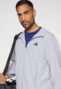 Ανοιχτό γκρι μπουφάν με φερμουάρ και μαύρο λογότυπο, με λεπτομέρειες από δίχτυ και ρυθμιζόμενη κουκούλα, φορεμένο πάνω από μπλε πουκάμισο, με τσάντα κρεμασμένη στον ώμο.