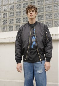 Sort bomberjakke med guld lynlås, ribbede manchetter, sidelommer og applikation. Båret over en sort T-shirt med blåt sommerfuglegrafik og slidte jeans.