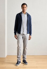 Un sweat à capuche zippé bleu marine sur une chemise à rayures, un jean gris clair et des baskets sombres avec des accents bleus. Sol en bois et fond neutre.