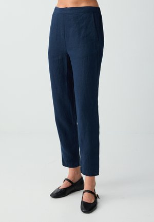 Donna con pantaloni dritti blu navy alla caviglia e ballerine nere Mary Jane, visibile dalla vita ai piedi su sfondo bianco.