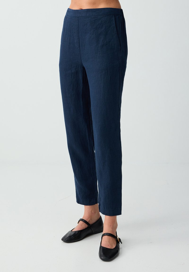 Femme portant un pantalon droit bleu marine longueur cheville et des ballerines Mary Jane noires, vue de la taille aux pieds sur fond blanc.