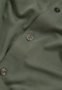 Țesătură de culoare verde măslinie cu un finisaj texturat, având butoane metalice rotunde și o cădere netedă. Amplasarea butoanelor pe margine accentuează designul.